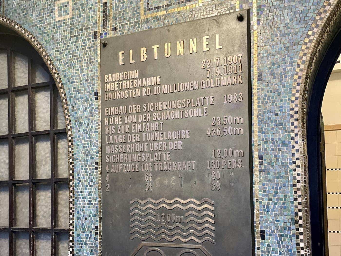 Metal plaque with embossed German text detailing the construction dates, costs, dimensions, and elevator capacities of Hamburg’s Old Elbe Tunnel, mounted on a mosaic-tiled wall.