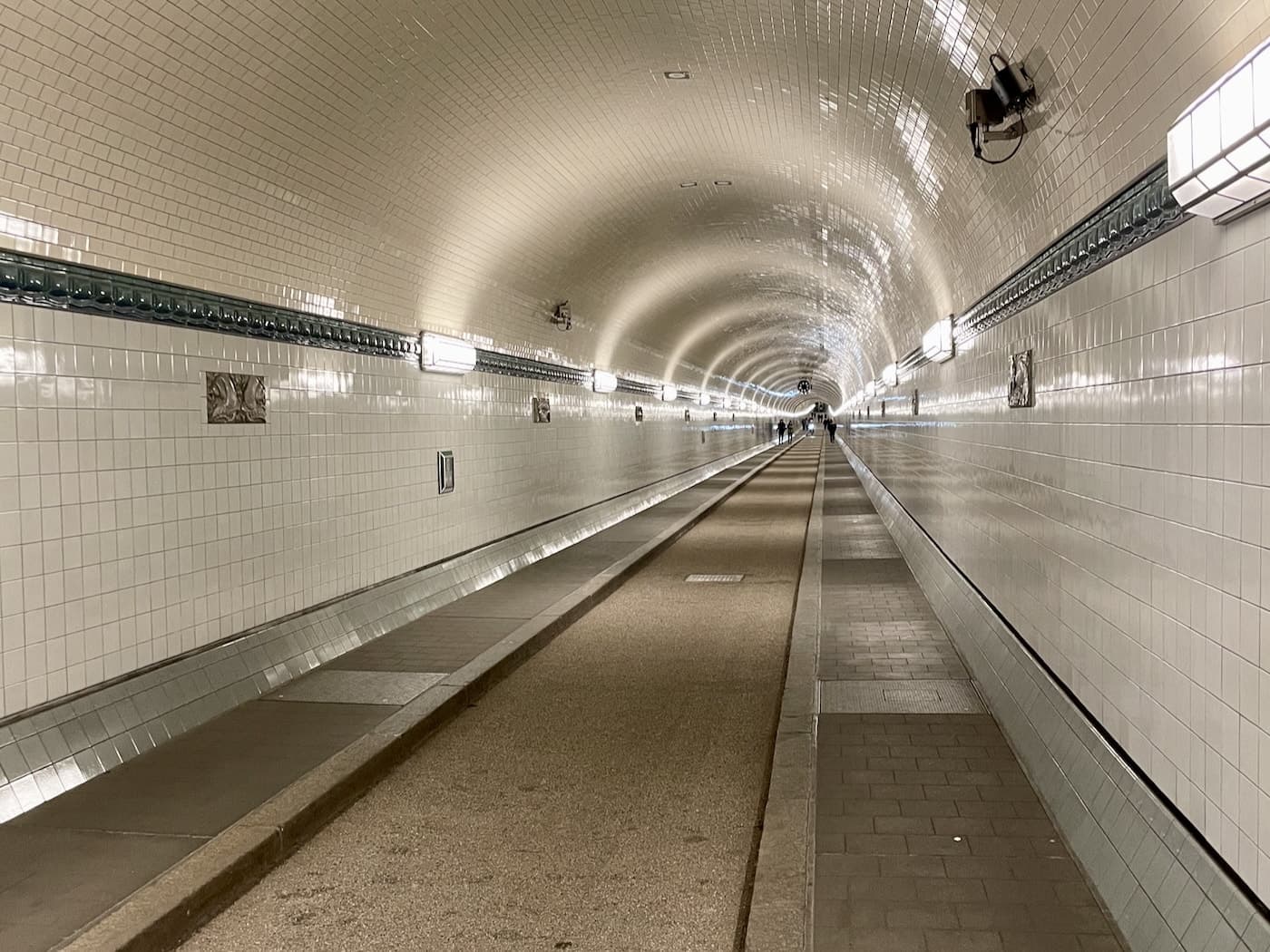 View down the tiled pedestrian tunnel under Hamburg's Elbe river, with curved ceiling, white and blue tiles, and evenly spaced rectangular light fixtures lining both sides