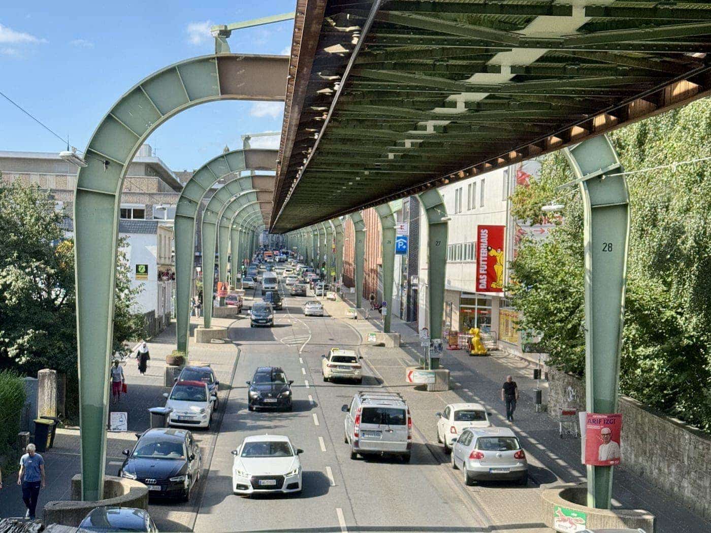 Suspended monorail track running above a busy urban street in Wuppertal, Germany, supported by giant green metal arches—an industrial-era marvel in a 2025 setting.