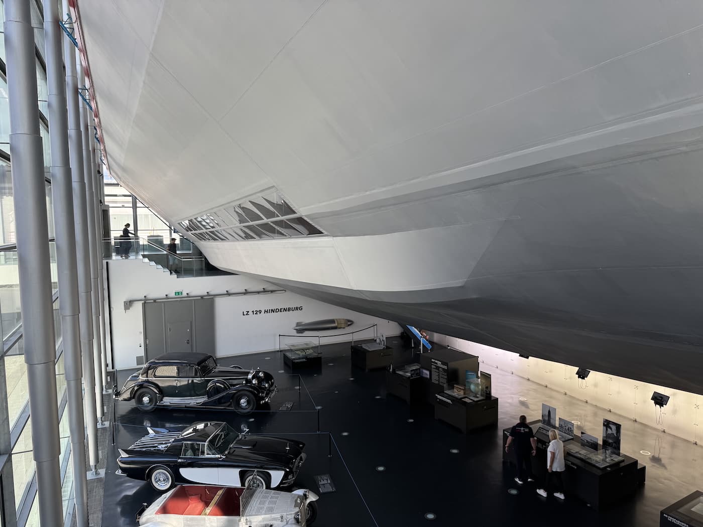 Indoor museum exhibit featuring a suspended model of part of the LZ129 Hindenburg airship above vintage automobiles and display cases.
