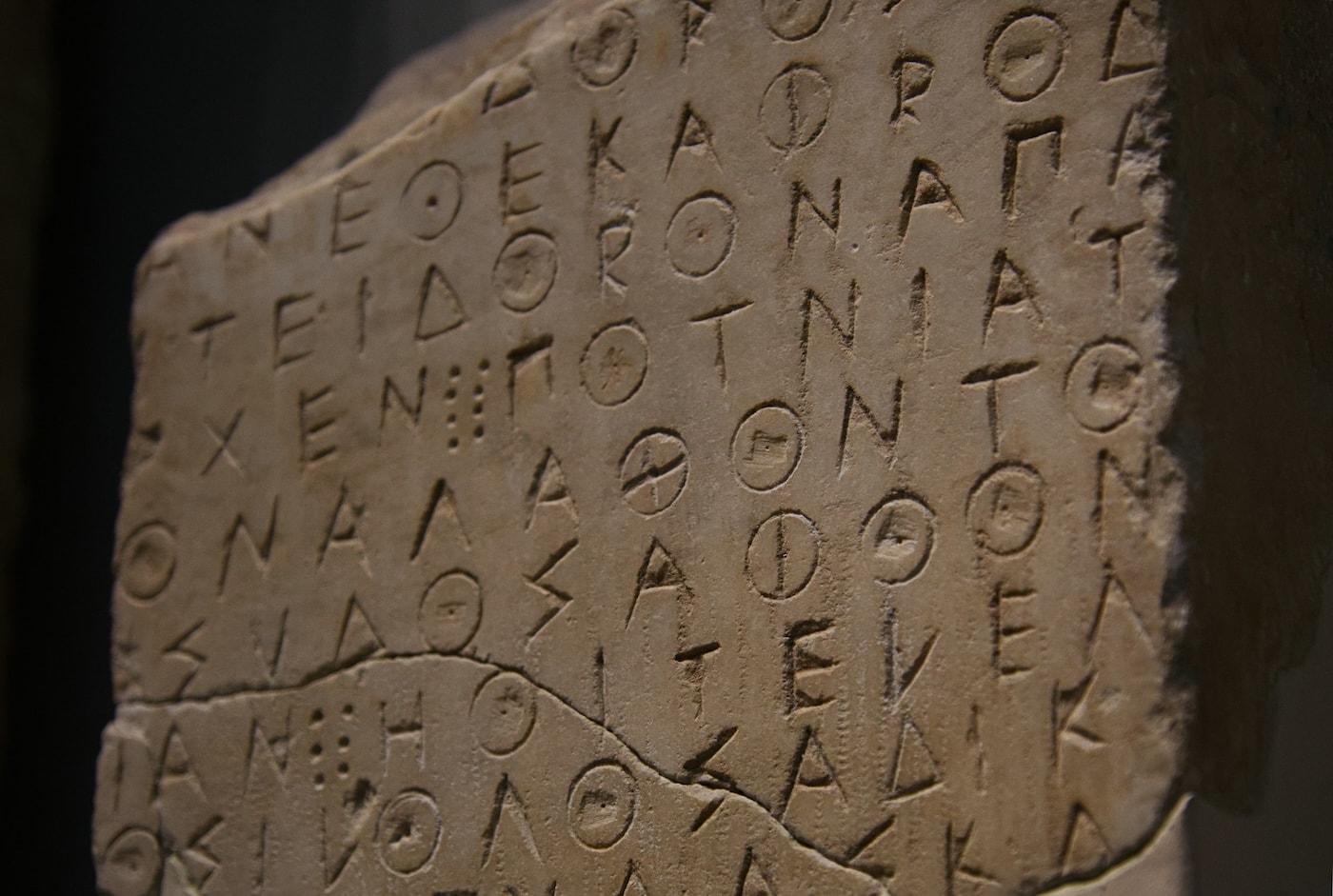 The angular carved shapes of Greek script line a shadowy ancient stone tablet set against a black background